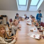 Skupina otrok sedi za mizo, pred njimi so krožniki in zavite sladkarije.