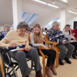 Več oseb različnih starosti sedi v vrsti in izpolnjuje liste s številkami.