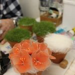 Na mizi stojijo majhni lončki z ročno izdelanimi cvetovi iz prosojnega materiala; ob njih so pripomočki za ustvarjanje in kozarci z vodo.