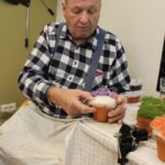 Starejši gospod sedi za mizo in izdeluje okrasni cvet v lončku; v rokah drži bel okrasni material ter natančno oblikuje aranžma.