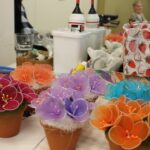 Na mizi je več pisanih cvetličnih aranžmajev v majhnih lončkih; v ozadju so škatle z materialom in ustvarjalni pripomočki.