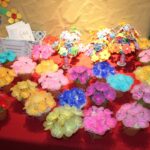 Na rdečem prtu je razstavljenih veliko barvitih ročno izdelanih cvetov v lončkih ter voščilnice in dekorativni izdelki.