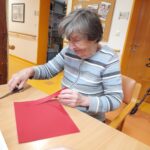 Ženska sedi za mizo in s škarjami reže rdeč list papirja.