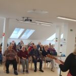 Stanovalci sedijo na stolih in vozičkih ter poslušajo glasbeni nastop; pogled je usmerjen proti izvajalkama.