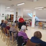 V sredini prostora stoji starejša gospa in govori zbranim stanovalcem, ki sedijo v polkrogu.