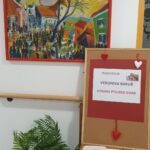 Na steni visi barvita slika mestnega prizora; ob njej je razstavna tabla z napisom »Razstavlja Veronika Rakuš – Utrinki Ptujske Gore« in odprta knjiga vtisov na mizici.