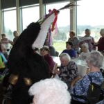 Oseba v črnem kosmatem pustnem kostumu stoji med sedečimi osebami v notranjem prostoru, okrašenem z visečimi barvnimi okraski.