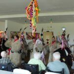 Več oseb v kosmatih pustnih kostumih stoji pred sedečimi osebami v prostoru z mizami in visečim barvnim okrasom.