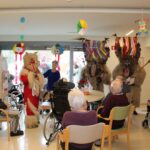 Več oseb v pustnih kostumih stoji v prostoru, medtem ko sedeče osebe opazujejo dogajanje okoli okrogle mize.