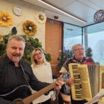 Tri osebe pojejo in igrajo glasbila, ena oseba drži kitaro, druga harmoniko.