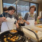 Dve osebi v predpasnikih pobirata ocvrto testo iz olja in ga odlagata v plastično posodo, v ozadju sedijo druge osebe.