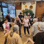 Skupina oseb stoji v krogu in se drži za roke, v ozadju glasbeniki igrajo kitaro in harmoniko.