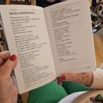 Oseba drži odprto knjigo z besedilom in jo bere, v ozadju sedijo druge osebe.