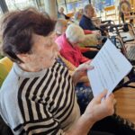 Ženska sedi na stolu in bere z lista papirja, medtem ko druge osebe sedijo v vrsti in poslušajo.