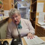 Ženska govori v mikrofon za pultom in bere iz odprte knjige.