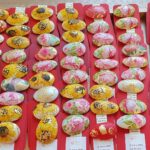 Na mizi so razporejeni pobarvani kamni z motivi cvetlic, pritrjeni na rdeče kartončke.