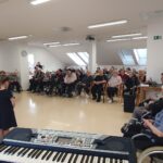 V svetlem prostoru sedi skupina oseb na stolih in invalidskih vozičkih, pred njimi stojijo otroci, v ospredju je električna klaviatura.