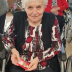 Oseba sedi na invalidskem vozičku in v rokah drži majhen rdeč kartonček z okrašenim kamnom.