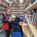 V knjižnici med policami s knjigami stoji nasmejana ženska s knjigo, ob njej pa moški sedi za računalnikom.