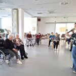 Skupina starejših sedi v krogu v svetlem prostoru, pred njimi stoji ženska z mikrofonom in govori.