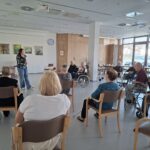 Udeleženci sedijo na stolih in poslušajo žensko, ki stoji spredaj in vodi dejavnost.