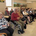 Skupina starejših sedi v prostoru in pozorno spremlja dogajanje.