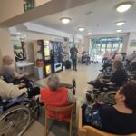 Udeleženci sedijo v krogu, pred njimi stoji ženska z mikrofonom in vodi srečanje.