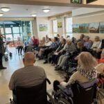 Večja skupina starejših sedi in posluša žensko, ki govori v prostoru z okni in slikami na stenah.