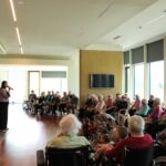 Oseba stoji in govori v mikrofon, pred njo sedijo osebe in otroci, ki jo poslušajo v svetlem prostoru.