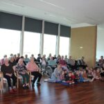 Oseba govori v mikrofon, pred njo na tleh sedi skupina otrok, okoli pa sedijo osebe na stolih in invalidskih vozičkih.