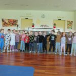 Skupina otrok stoji v vrsti in drži drug drugega za ramena, ob njih sedita dve odrasli osebi.