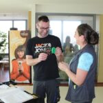 Moški drži zelen predmet v obliki kaktusa in ga kaže ženski, ki stoji nasproti njega. Oba se smejita in gledata predmet. V ozadju je stojalo z reklamnim plakatom in miza z materiali.