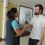 Ženska drži zelen predmet v obliki kaktusa in ga izroča moškemu, ki stoji nasproti nje v hodniku. Oba se pogovarjata in se gledata. V ozadju je stena z obešenim plakatom in okvirjeno sliko.