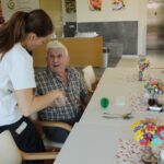 Ženska se nagne k moškemu, ki sedi za mizo v jedilnici, in mu nekaj pokaže ali izroči. Na mizi je zelen predmet v obliki kaktusa, šopek pisanih cvetov iz papirja in pribor. Nasproti sedi druga oseba.