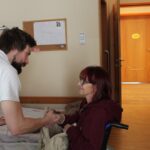 Moški stoji ob postelji in se rokuje z žensko, ki sedi na stolu v sobi.