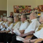 Skupina oseb sedi v vrsti, drži liste papirja in sodeluje pri skupnem petju.