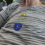 Na vratu osebe je vidna ogrlica z modrim cvetličnim obeskom.