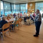 Moški stoji v prostoru in bere z lista papirja, medtem ko več oseb sedi za mizami in posluša.