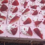 Kosi rožnate sladice so razrezani na kvadrate in okrašeni s stepeno smetano ter koščki jagod.