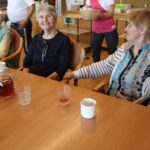 Tri ženske sedijo za mizo s skodelicami in vrčem pijače, v ozadju oseba prinaša krožnike.