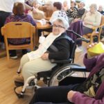 Več oseb sedi za mizami in pije iz skodelic, v ospredju oseba na invalidskem vozičku opazuje dogajanje.