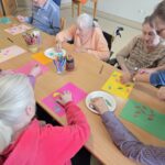 Osebe sedijo za mizo in iz barvnega papirja ter flomastrov izdelujejo likovne izdelke z motivi listov.