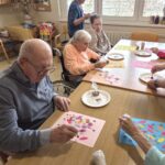 Osebe sedijo za mizo in s pomočjo barv ter štampiljk ustvarjajo cvetlične motive na papirju.