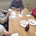 Osebe sedijo za mizo in z barvicami rišejo rastlinske motive na manjše liste papirja.