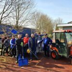 Skupina oseb stoji na prostem ob prikolici in majhnem traktorju ter drži orodje za grabljenje in čiščenje listja.
