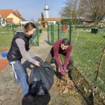 Oseba zbira suho listje ob ograji in ga podaja drugi osebi, ki drži odprto vrečo za odpadke.