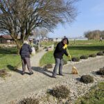 Dve osebi pometata tlakovan pot v parku, v ozadju so drevesa, klopi in travnata površina.