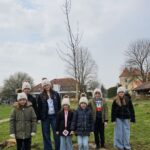 Skupina oseb stoji na travniku ob na novo posajenem drevesu in gleda proti kameri.
