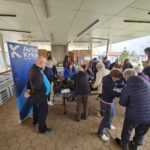 Več oseb stoji okoli mize pod nadstreškom in skupaj presaja rastline v lonce.