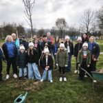 Skupina oseb stoji na travniku ob drevesu in vrtnarskem orodju ter pozira za fotografijo.